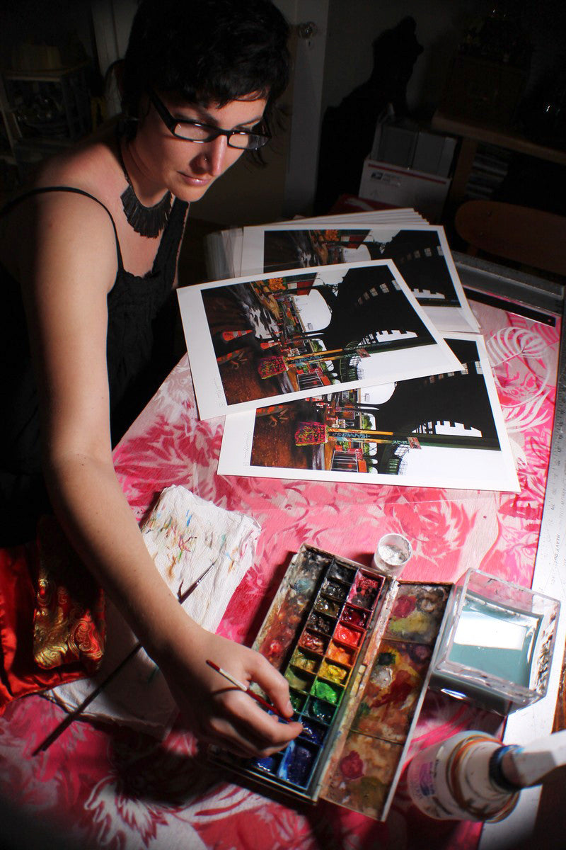 A person with short dark hair and glasses paints at a table covered in a red patterned cloth, surrounded by cityscape prints. They use archival Epson Ultrachrome HDR inks from the Jessica Hess watercolor set, enhancing the artistic and focused atmosphere.