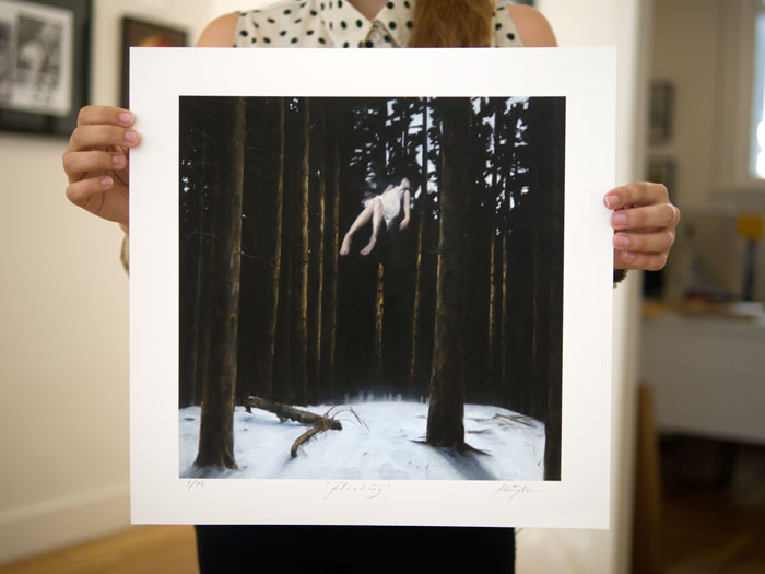 A person in a sleeveless polka dot shirt holds a bordered fine art giclee print by Henrik Uldalen, showcasing a levitating figure in a dark, snowy forest. The rooms art-adorned walls amplify the prints ethereal charm.