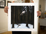 A person in a sleeveless polka dot shirt holds a bordered fine art giclee print by Henrik Uldalen, showcasing a levitating figure in a dark, snowy forest. The rooms art-adorned walls amplify the prints ethereal charm.