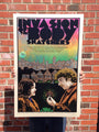 A person showcases a vibrant Chuck Sperry poster for Invasion of the Body Snatchers on a brick wall, featuring two figures, a cityscape with houses under a starry sky, and bold purple and green text.