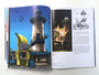 An open hardcover book showcases an illustrated lighthouse. The left page displays a lighthouse with a large banana sign and birds, akin to Tim Doyle’s UnReal Estate style, while the right page features text and dramatic images, including one of the lighthouse engulfed in flames.
