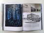An open hardcover book shows two pages: the left with a forest scene illustration in dark blue and the right with a grayscale image of a Cinema building, showcasing UnReal Estate II, inspired by Tim Doyles art.