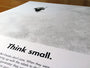 A detailed view of a minimalist design printed page showcases a small helmet image at the top. The bold text says, Think small. Its an NYCC 2015 Exclusive from Justin Van Genderens limited edition screen print collection, with some obscured text at the bottom.
