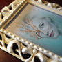 An ivory frame displays an artwork of a womans face blending realism and abstraction, reflecting Bec Winnels style. Delicate orange and blue pencil details extend from her hair, with half of her features realistic and the other abstract.