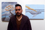 A short-haired, bearded person stands before vibrant paintings on a white wall. The left features swirling branches on wood by Roland Tamayo, while the right shows a whale with a bridge and orange trail, suggesting art for sale.