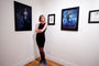 A person in a black dress smiles with crossed arms in an art gallery by a wall featuring framed Allison Reimold artworks, including Ritual Study in graphite and gouache, alongside images of a person with birds and a portrait. The floor is wooden.