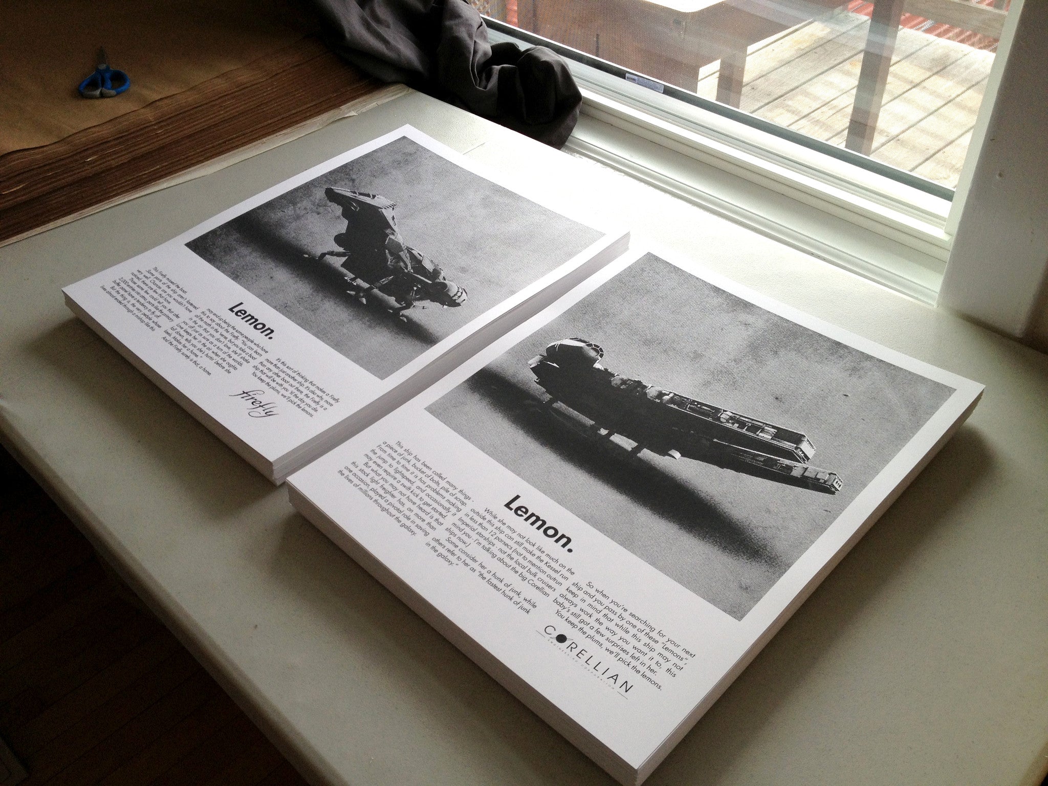 Two limited edition posters by Justin Van Genderen on a table by the window showcase spaceships titled Lemon. The vintage car ad-inspired designs feature one cylindrical and one disc-like ship. Scissors nearby hint at the precision of screen print artistry.