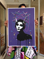 A person displays a vibrant Eelus limited edition print of a stylized womans face with dripping paint, antlers, and colorful strings. The purple background enhances her pop art skirt.