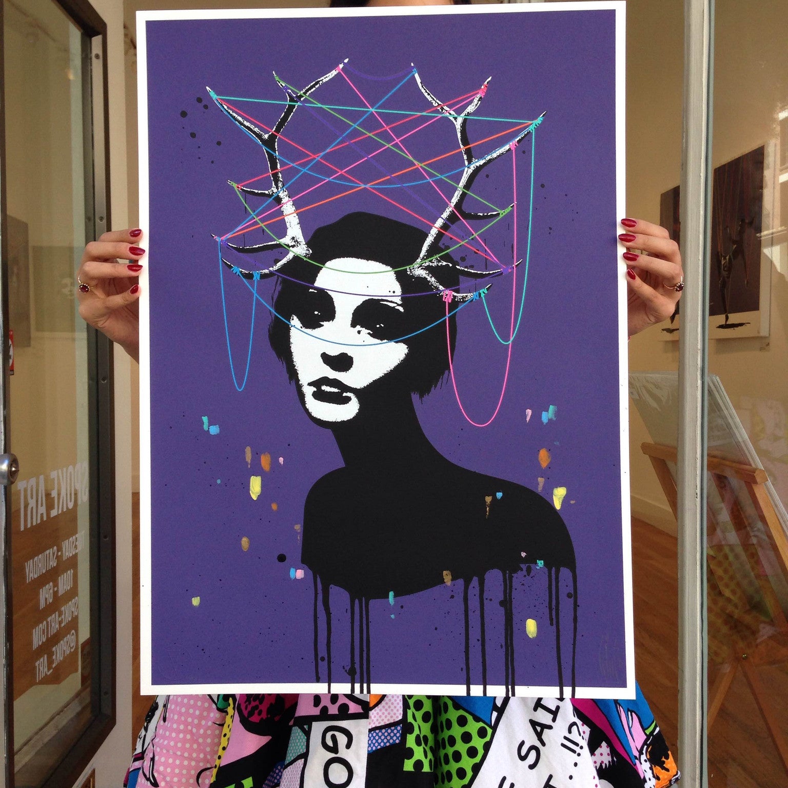 A person displays a vibrant Eelus limited edition print of a stylized womans face with dripping paint, antlers, and colorful strings. The purple background enhances her pop art skirt.