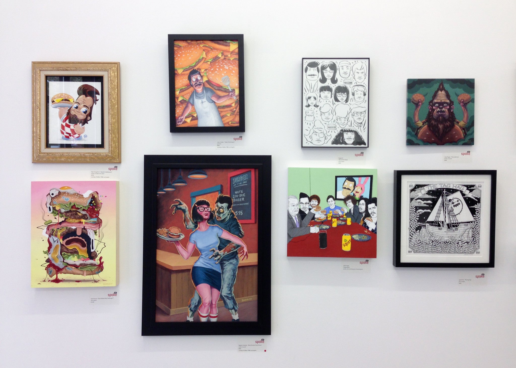 A collection of framed artworks by Ryan Hungerford on a white wall showcases vibrant, abstract, and cartoonish illustrations. Inspired by the playful vibe of a Bobs Burgers art show at Spoke Art NYC, each piece—from whimsical portraits to quirky depictions of food and Big Bobs BeefSquatch—is distinct.