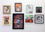 A collection of framed artworks by Ryan Hungerford on a white wall showcases vibrant, abstract, and cartoonish illustrations. Inspired by the playful vibe of a Bobs Burgers art show at Spoke Art NYC, each piece—from whimsical portraits to quirky depictions of food and Big Bobs BeefSquatch—is distinct.