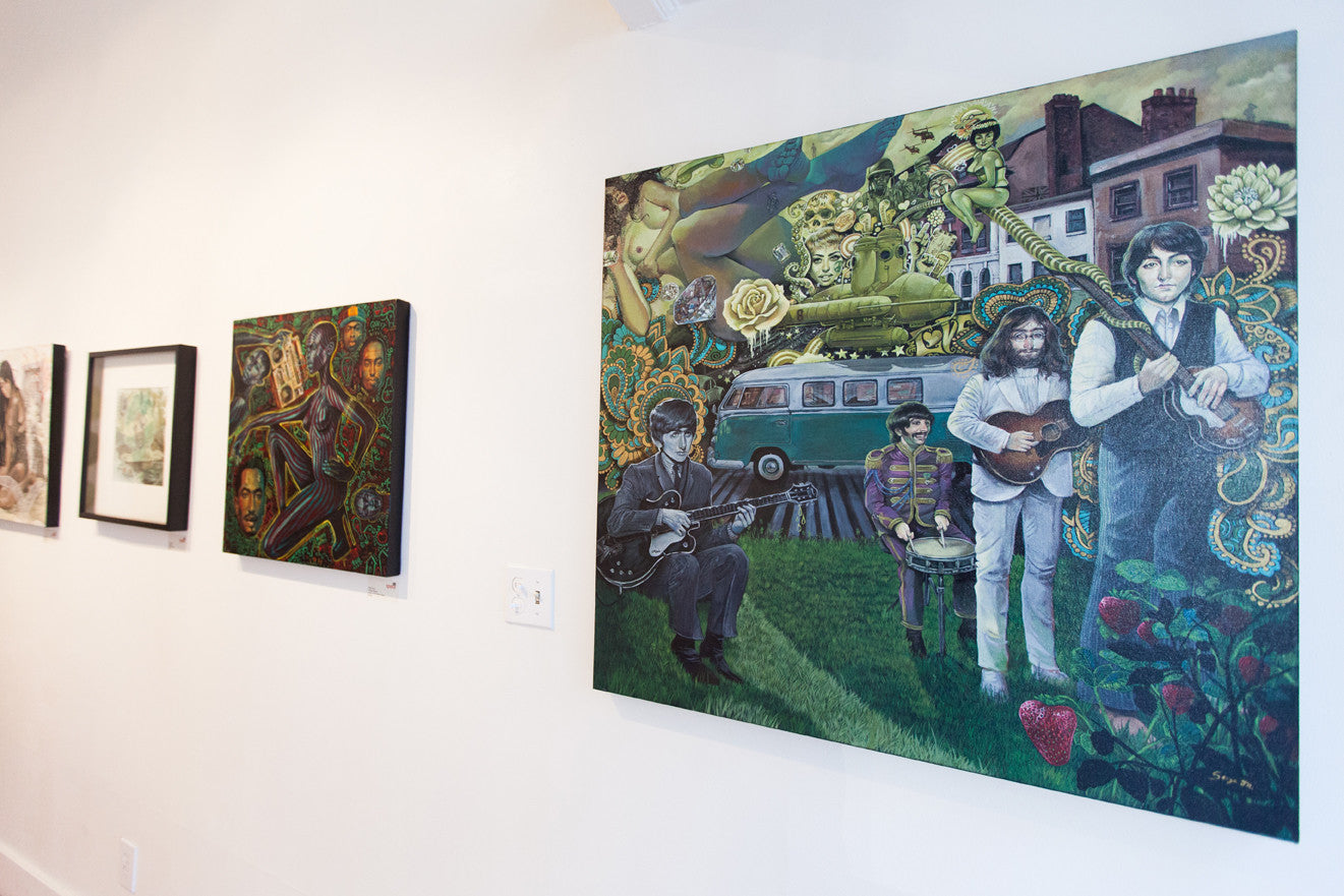 A gallery wall features several artworks, highlighting a Serge Gay Jr. painting of four musicians in a surreal, colorful scene with a bus and roses. Smaller pieces, such as an Abstract Rhymes canvas, hang on the left. The room retains a bright, minimalist feel.