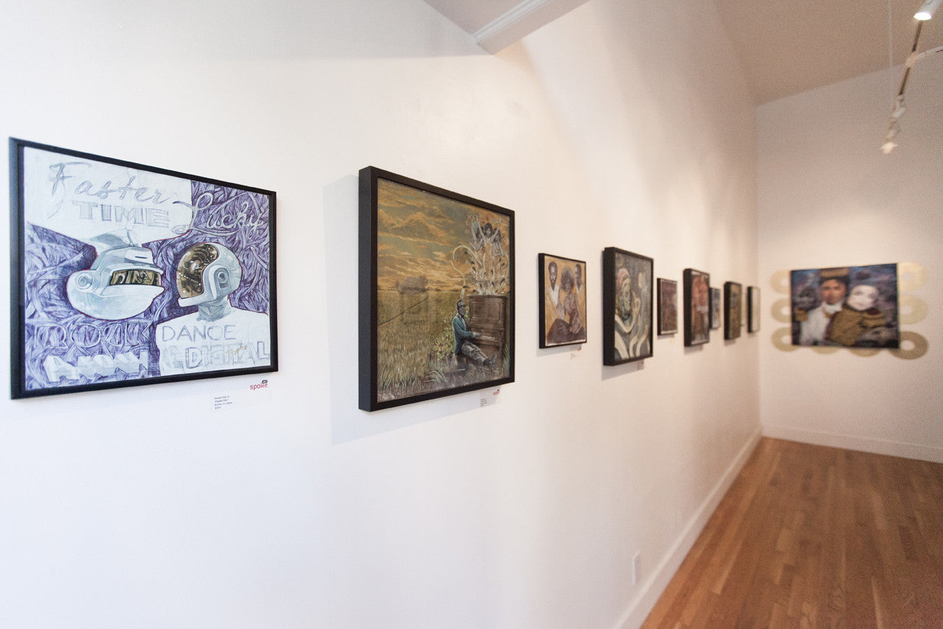 A gallery hallway features Serge Gay Jr.s vibrant framed artwork on an off-white wall. The largest painting at the end depicts two people. The wooden floor shines brightly, casting a glow reminiscent of Digital Star across various subjects and styles.