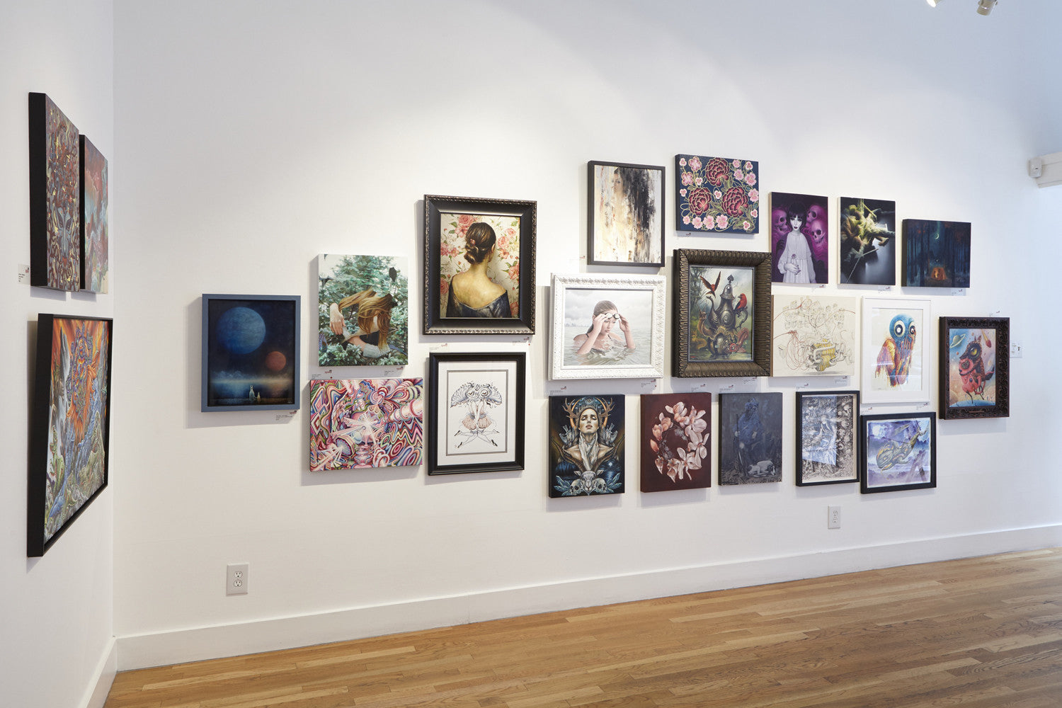 A gallery wall featuring diverse framed artworks, such as portraits, abstracts, and colorful illustrations by Meghan Howland, adorns a white wall above a wooden floor.