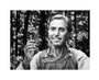 A grayscale image of a man outdoors, wearing a buttoned shirt under overalls and holding a small comb in his right hand while smiling slightly. Blurred trees suggest a forest setting. This Joel Phillips archival pigment print comes custom framed to enhance its charm.