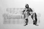 A man in a hooded jacket and baggy pants relaxes on a partially sketched couch. Rendered with charcoal and graphite, the black-and-white background contrasts the grayscale details of the man. A book or magazine lies beside him, echoing Joel Phillips art seen at San Francisco fairs.