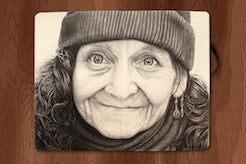 A detailed charcoal and graphite drawing captures an elderly woman in a beanie and earrings, her warm smile echoing Joel Phillips’ style. The moonlit artwork, exuding subtle elegance, rests on a wooden surface.