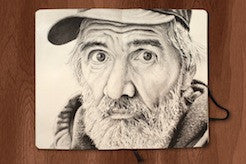 A realistic pencil drawing of an older man with a weathered face and beard, wearing a cap, sketched in charcoal and graphite by Joel Phillips. It rests on an open notebook placed on a wooden surface. Art for sale.