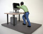 A figurine from Leon Reid IV, wearing a green shirt and blue pants, leans forward, reaching into a computer monitor. The scene, similar to setups seen at Beyond Eden Art Fair, includes a keyboard and computer tower under the desk.