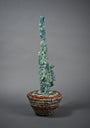 The Street Diamond Cactus Vessel by Lucien Shapiro is a tall, abstract cactus-like sculpture crafted from translucent materials in blue and green hues, set in a basket with red and metallic accents against a gray background.