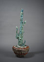 A tall, abstract sculpture in the style of Lucien Shapiros blue-green Street Diamond Cactus Vessel stands in a bowl with a textured red and brown design, set against a plain gray background.
