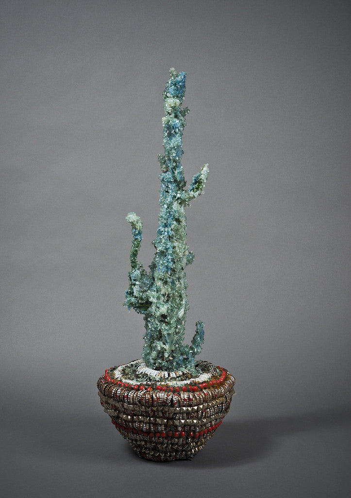 A tall, abstract sculpture in the style of Lucien Shapiros blue-green Street Diamond Cactus Vessel stands in a bowl with a textured red and brown design, set against a plain gray background.