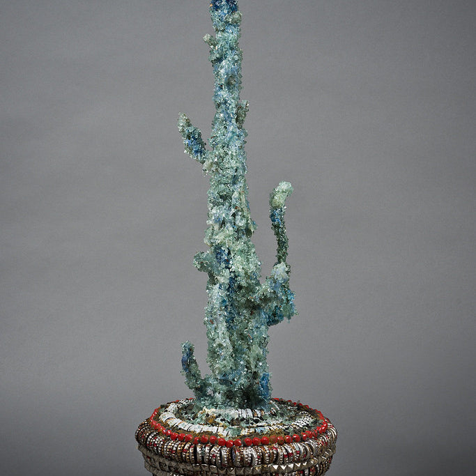 The Street Diamond Cactus Vessel by Lucien Shapiro is a tall, slender sculpture adorned with sparkling blue crystals. It stands in an intricately woven red and gray basket, set against a plain gray background, embodying Shapiro’s unique bottle cap art style.