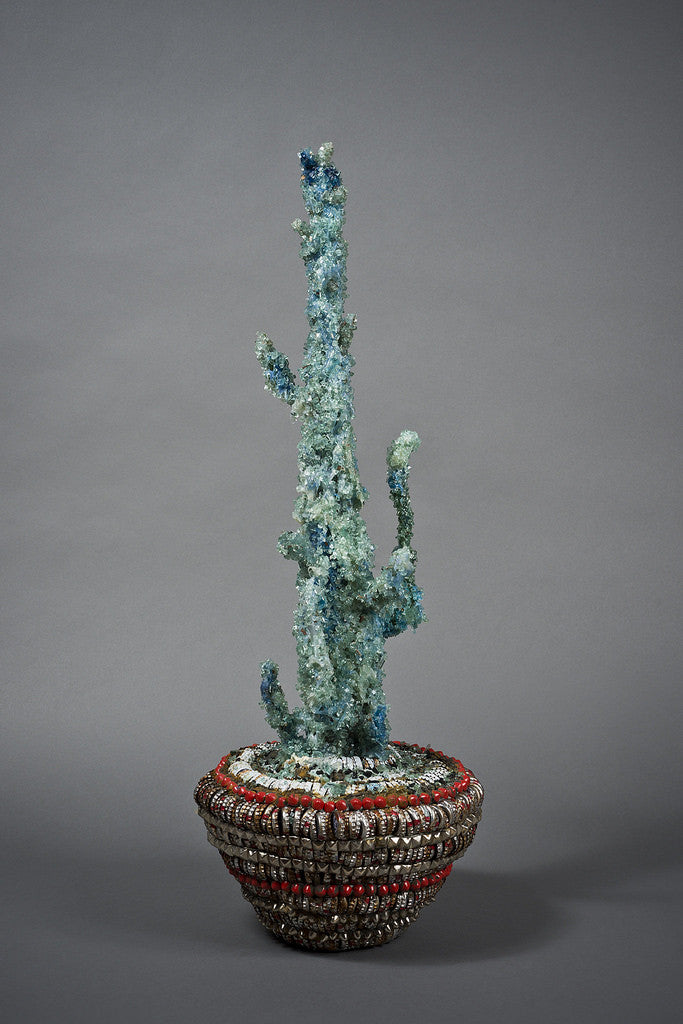 The Street Diamond Cactus Vessel by Lucien Shapiro is a tall, slender sculpture adorned with sparkling blue crystals. It stands in an intricately woven red and gray basket, set against a plain gray background, embodying Shapiro’s unique bottle cap art style.