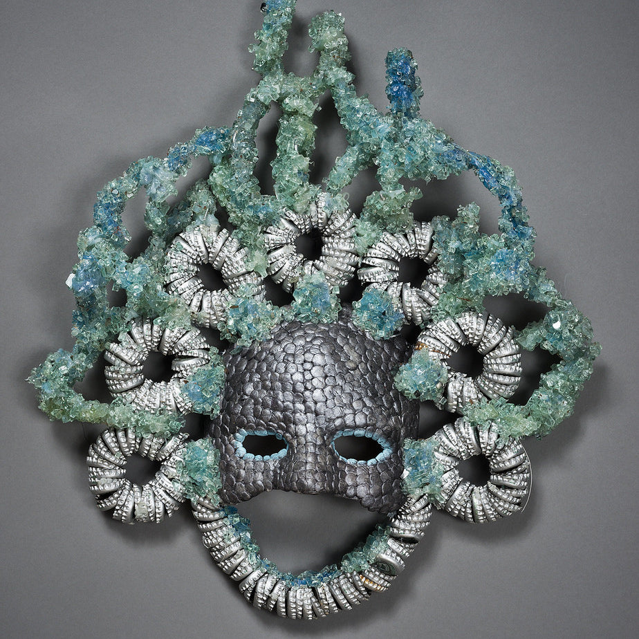 The Silver Streaks Mask by Lucien Shapiro has a textured, scaly surface like a human face, with curly metallic silver coils and blue-green crystals resembling hair. It includes eye and mouth openings.