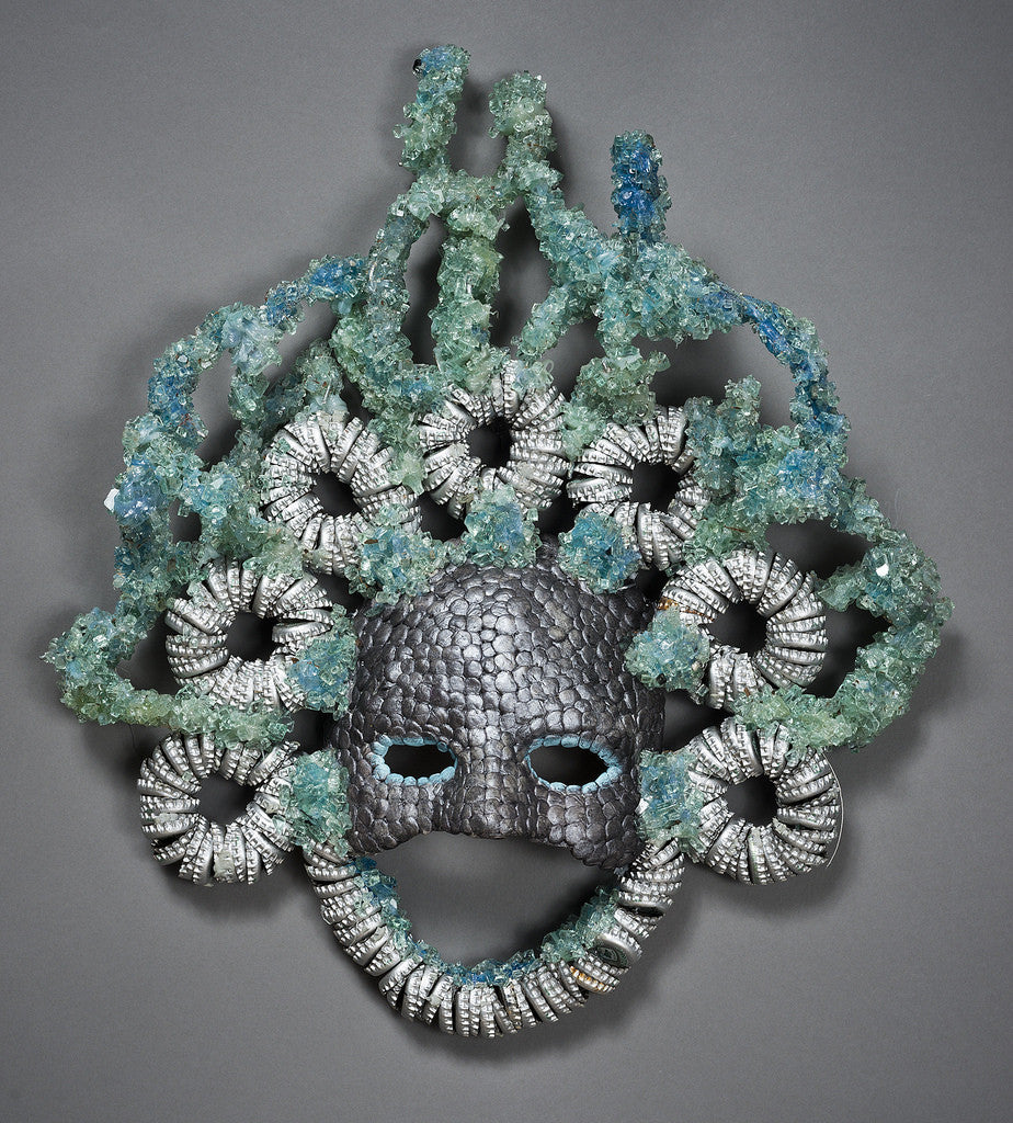 The Silver Streaks Mask by Lucien Shapiro has a textured, scaly surface like a human face, with curly metallic silver coils and blue-green crystals resembling hair. It includes eye and mouth openings.