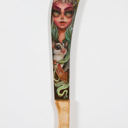 A Tatiana Suarez machete with a wooden handle features a mystical woman from Spoke Arts style. Inspired by the Mira oil series, she has green hair, a colorful headpiece, and holds a small mammal set against vibrant green and yellow hues.