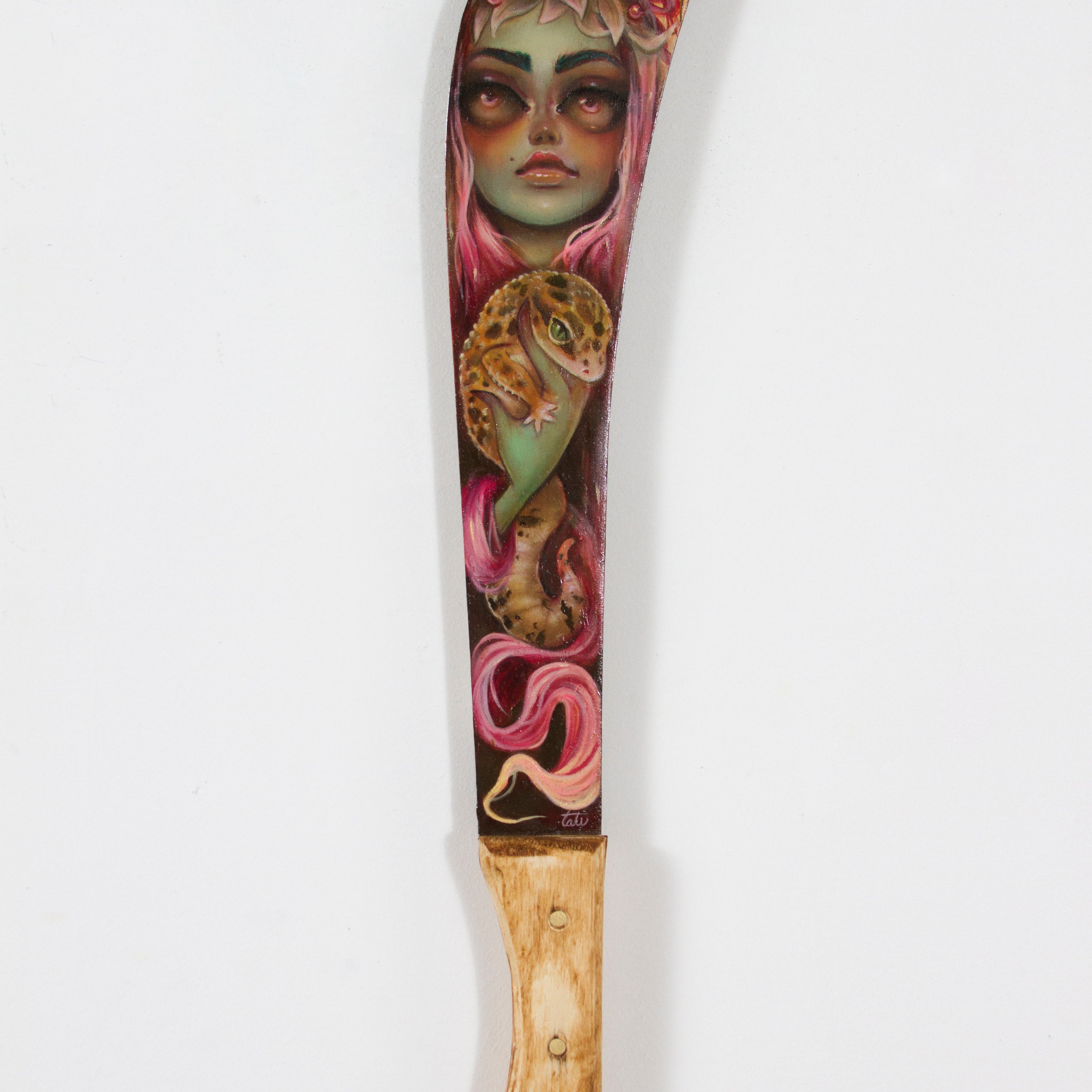 Tatiana Suarezs intricately painted machete at Spoke Art features a wooden handle and stunning blade design: a green-skinned woman with pink hair surrounded by leaves, butterflies, and a lizard, beautifully merging nature and fantasy elements.