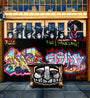 Graffiti artwork on a garage door features stylized faces and vibrant tags in a Jessica Hess style. A wooden table with a painted face is in front, set against brickwork, a large window, and a No Parking sign skillfully integrated into the graffiti scene.
