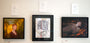 Three Barron Storey artworks on a wall: a portrait with a yellow backdrop, an abstract collage in ink and acrylic, and a painting of someone among autumn leaves. Each frame has a white card with text above it.
