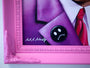 This limited edition framed image by Scott Scheidly showcases a person in a purple suit and a black button with a white sad face. The pink ornate frame, which is chipped at the corner, and the button are prominent features, along with the artist’s signature.