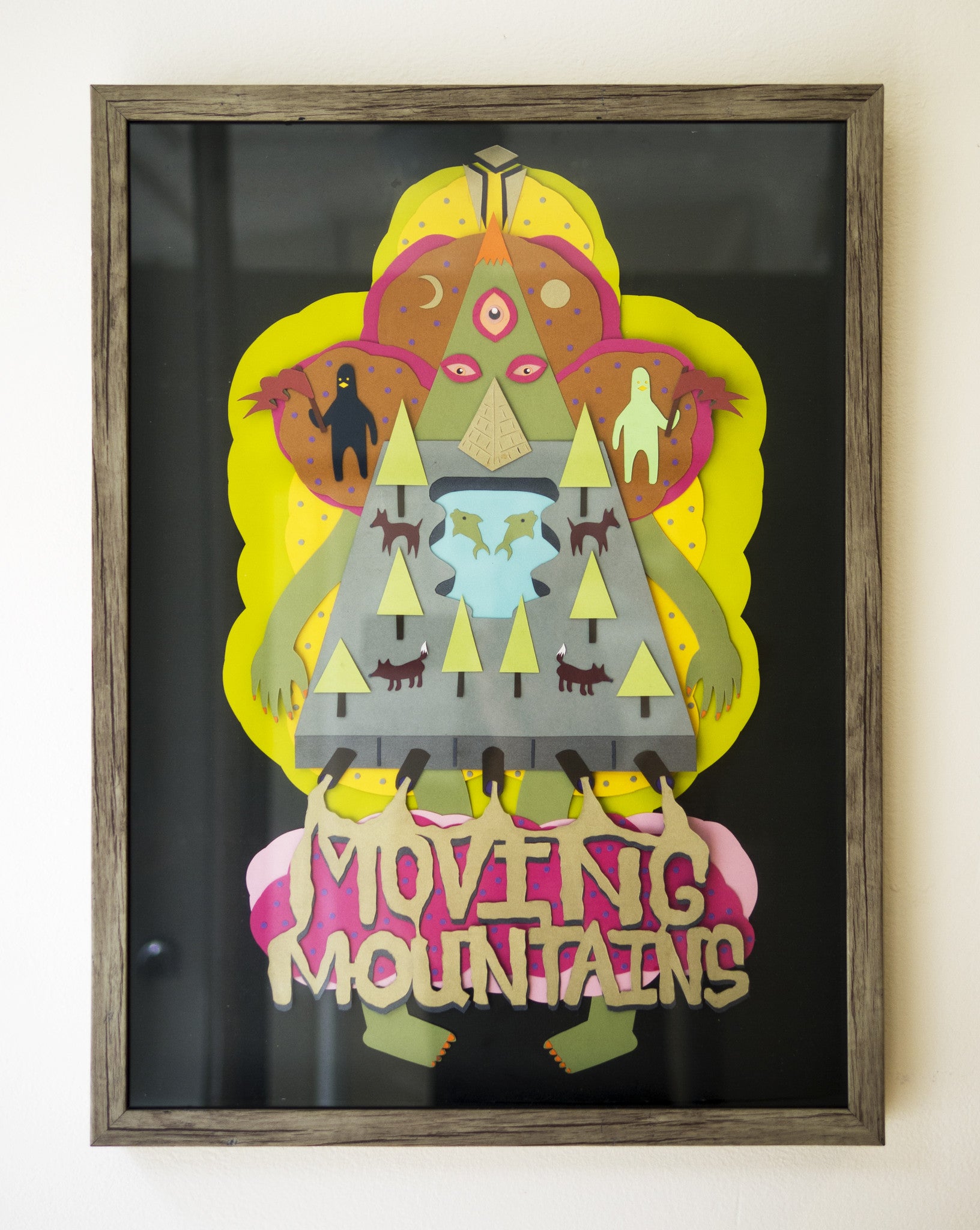 A framed artwork by Daniel Jaffe showcases a colorful figure with a triangular body, trees, animals in layered paper, and a multi-eyed face against vibrant backgrounds. The phrase MOVING MOUNTAINS is at the bottom.