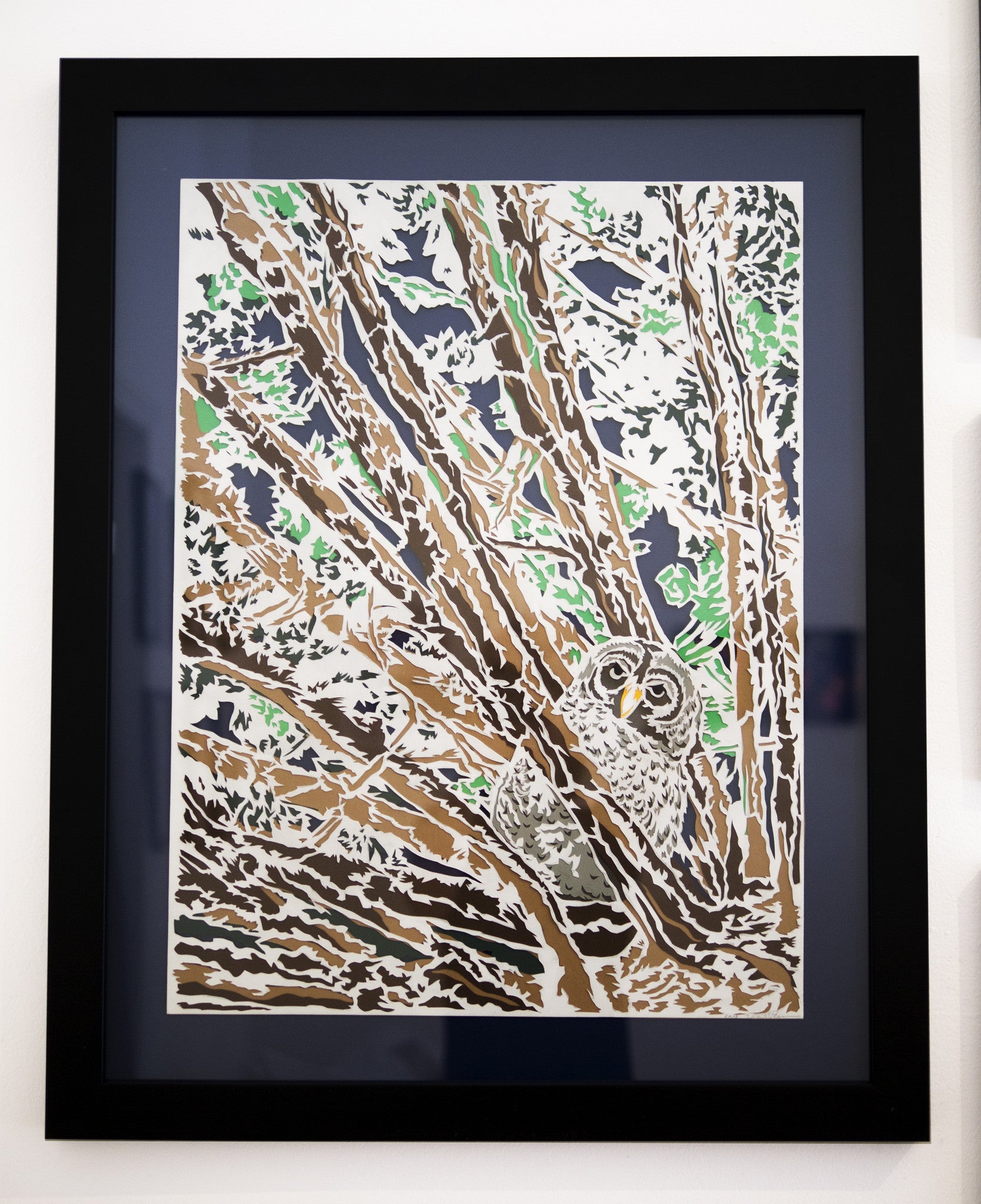 Framed artwork The Watcher by Elaine Penwell features an owl nestled in brown tree branches with sparse green leaves. Its large eyes stand out against a detailed, abstract background crafted from hand-cut paper.