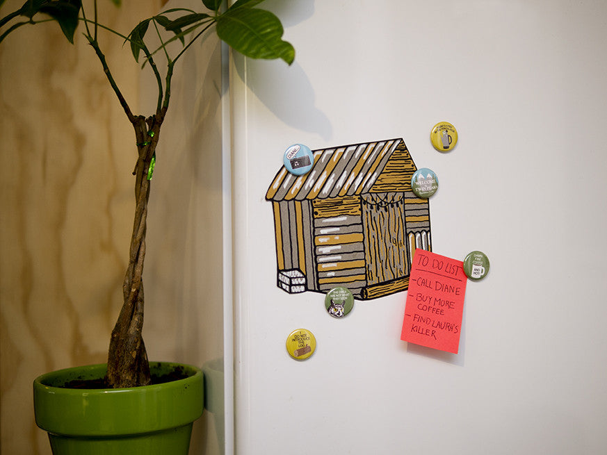 A small tree in a green pot sits by a white wall with a cartoon shed drawing. Scattered around are round magnets from Arthurs Plaid Pants Twin Peaks set, and theres also a pink sticky note with a to-do list.
