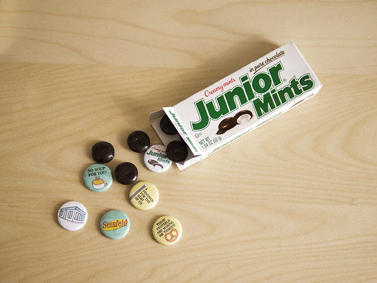 An open Junior Mints box spills its candies on a wooden surface. Nearby lie six 1 Seinfeld magnets by Arthurs Plaid Pants, featuring designs and texts like No soup for you and Serenity now.