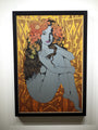 A framed Chuck Sperry artwork features a woman akin to Ariadne, seated with knees drawn up, and her hair adorned with bright orange flowers. The backdrop is an abstract yellow and orange pattern, beautifully presented on an oak panel.