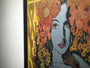 A framed artwork on an oak panel by Chuck Sperry shows Ariadne with purple lips, a blue face, and is surrounded by orange and yellow flowers. The background has abstract patterns in brown and yellow tones, emphasizing Sperrys distinctive style.