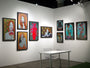 The art gallery wall features framed, colorful portraits of women, including Caia Koopmans works. A small white table with a transparent chair is set before vibrant paintings, some made with acrylic on wood panel, beautifully lit to highlight intricate details.