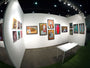 The gallery exhibits Caia Koopmans vibrant, abstract acrylic paintings on white walls. Her unique style is showcased on wood panels. A white table with a clear chair rests on the green carpeted floor, while overhead lighting highlights the colorful artwork.