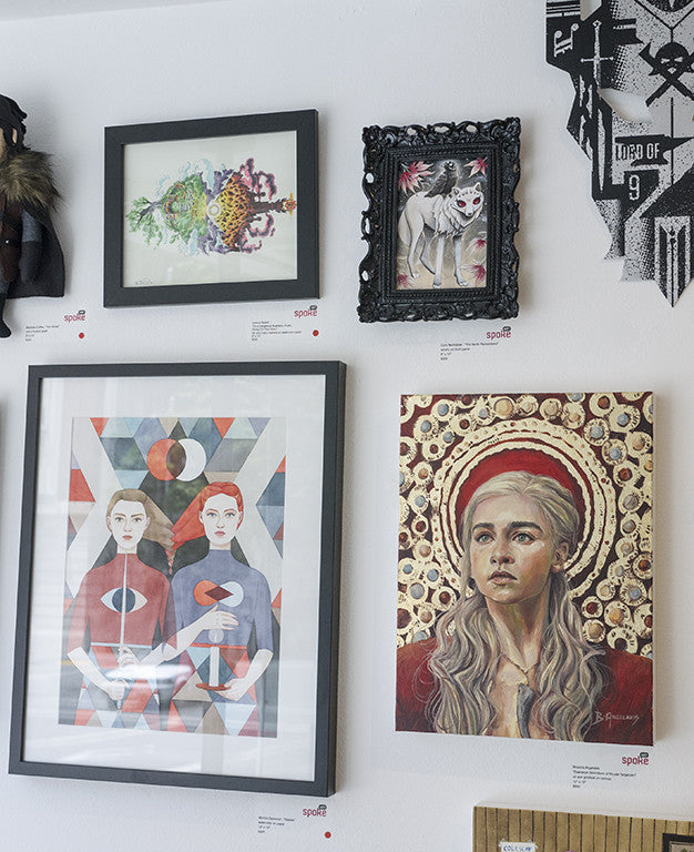 A gallery wall at Spoke Art features diverse artworks: a colorful abstract fish, a Joshua Budich-framed wolf, a geometric print of two people, a striking portrait of a woman with red halo, and other decorative pieces inspired by Lord of the Thrones.