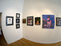 A gallery room showcases framed artworks by M Ian Dringenberg on white walls. The paintings and photos vary in style, with abstract and portrait elements. The wooden floor complements each piece, labeled with details such as Ink on paper.