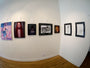 A gallery wall showcases framed artworks, featuring vibrant abstracts and portraits. Among them is Love Without Limits by M Ian Dringenberg. The space features wooden floors, white walls with title cards above each piece, well-lit and organized with graphite and charcoal accents.