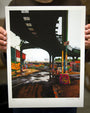 A person displays a vibrant Jessica Hess urban artwork on cold press watercolor paper, depicting a rainy street beneath an elevated train. Bright colors and puddle reflections enrich the scene, adding depth to the industrial setting.