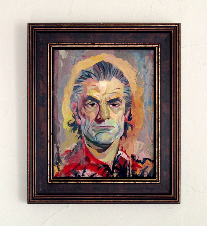 A vibrant portrait by Rich Pellegrino depicts an older man with gray hair and expressive, serious features. Elegantly framed in dark wood, it is showcased on a white textured wall at the Spoke Art gallery.