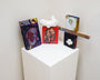 A white pedestal showcases four bold abstract paintings, with two small sculptures incorporated: one titled On the Water atop the red painting and another on a wooden stick, evoking Tessar Los style.