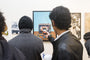 At an art gallery, a group admires Allan Innmans oil on canvas of a retro TV with an eye. Someone in a leather jacket takes a photo of the artwork, while another sips from a cup discussing art sales amidst various pieces on the walls.